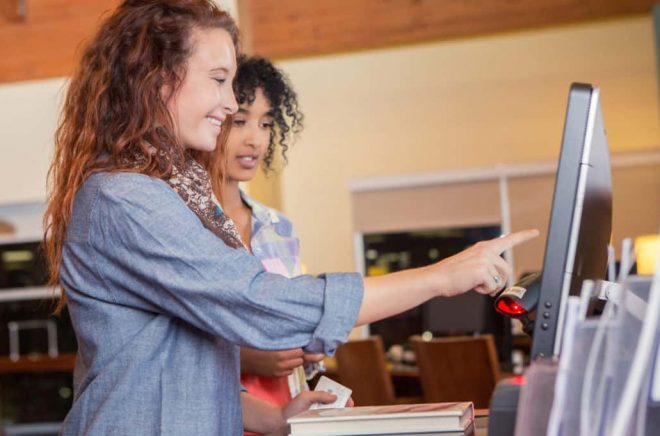 Caucasian and African American young women are patrons in modern public library. Students or customers are standing in line, using touch screen self check out kiosk to borrow books. Young women are wearing trendy clothing.