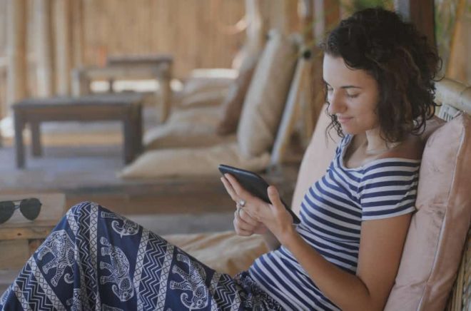 Cute tanned girl in a striped T-shirt and blue pants sitting outdoor on a chair and reading a romantic story on the ellektronnoy book with a smile.