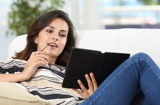 Portrait of a happy woman reading a book in an ebook sitting on a sofa at home