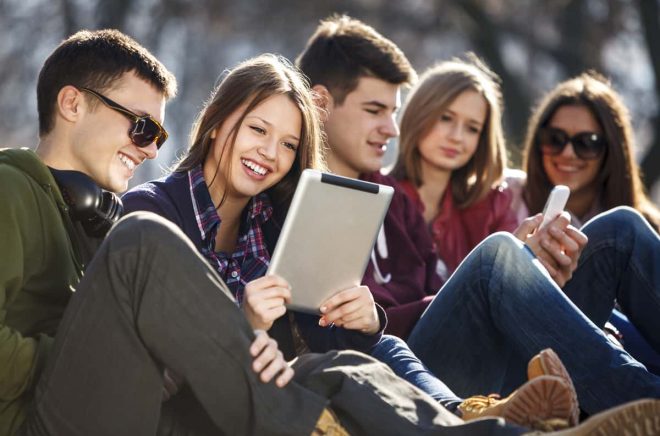 A group of young people, teenagers  are smiling and having fun outdoors. The group consists of two boys and three girls. A boy in a green jacket wearing sunglasses and a girl with brown hair are having fun while watching something on a digital tablet. Another boy is holding and watching something on a mobile phone with other two girls.