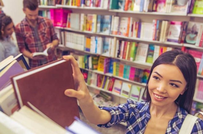 High angle view of attractive girl smiling while choosing book at the bookshop, young people in the background