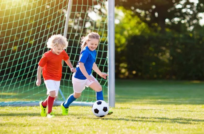 Kids play football on outdoor field. Children score a goal at soccer game. Girl and boy kicking ball. Running child in team jersey and cleats. School football club. Sports training for young player.