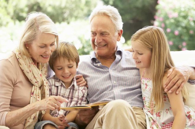 Portrait Of Grandparents Reading To Grandchildren On Sofa Smiling